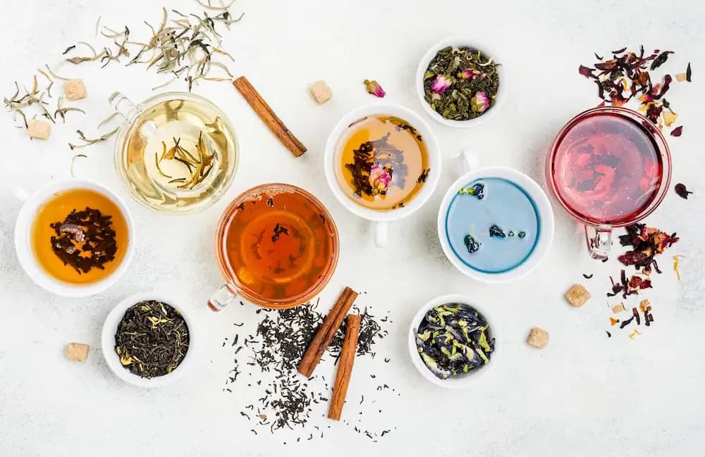 A collection of six tea cups with teas inside them surrounded by different herbs